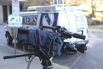 A video camera pointed to the side is shown in front of a white and blue news van