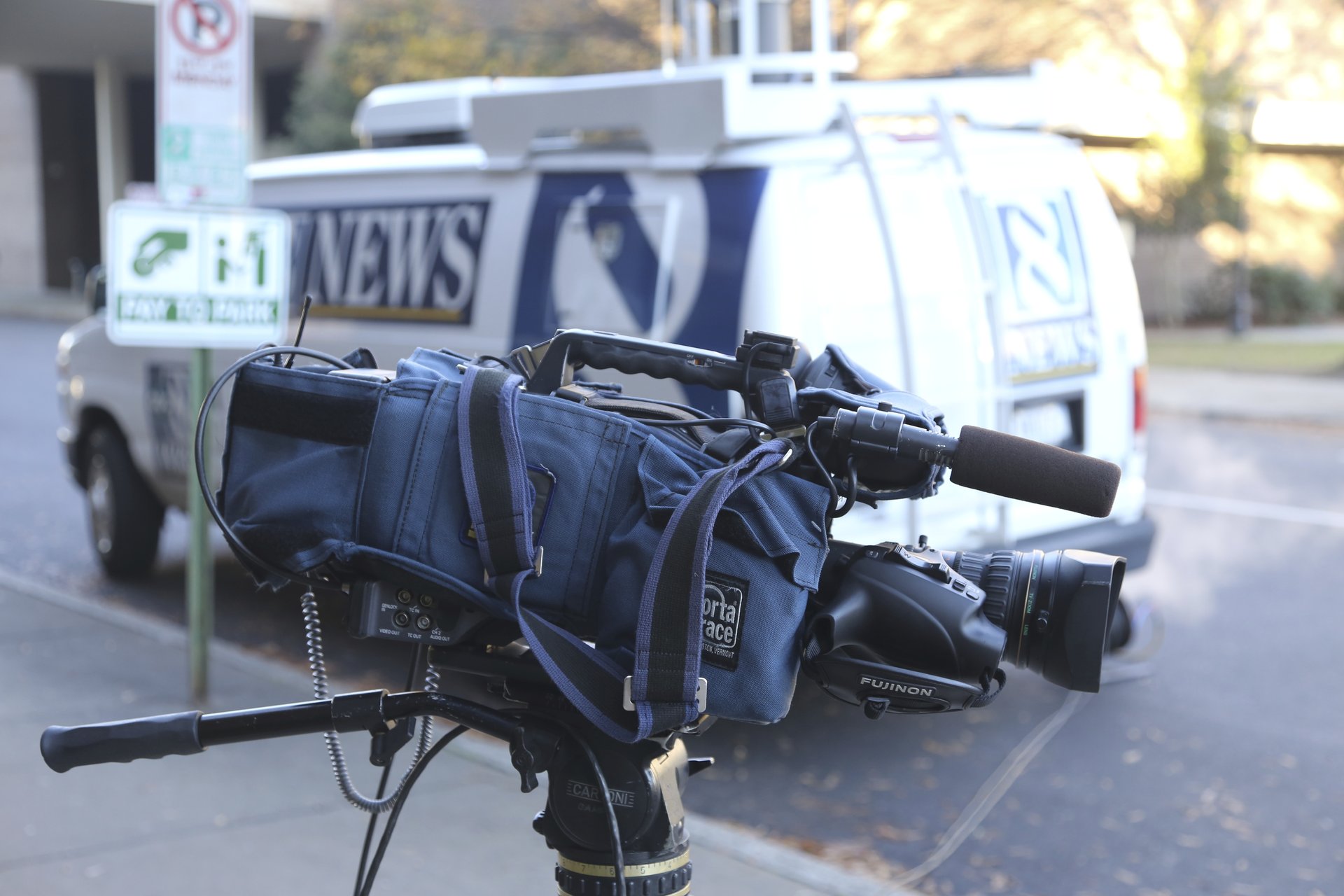 A video camera pointed to the side is shown in front of a white and blue news van