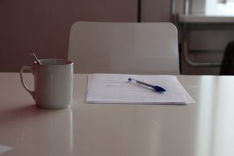 A small pile of papers and a pen on top of a desk, and a mug alongside it.