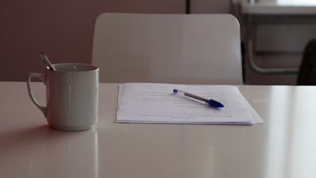 A small pile of papers and a pen on top of a desk, and a mug alongside it.