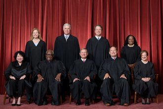All of the U.S. Supreme Court Justices pose for an official portrait in their black robes before a red curtain. Five justices sit and four stand behind them.