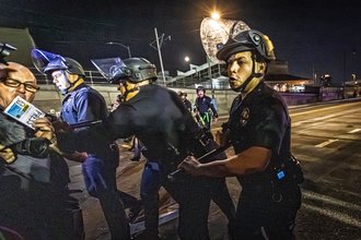 Night time image of Nick Stern, left, standing on a street and holding a camera and a badge that says PRESS. In the center are three police officers advance on Stern, holding police batons. One officer is pushing Stern.
