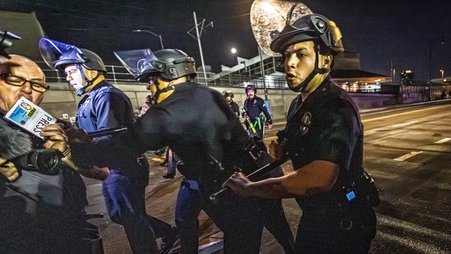 Night time image of Nick Stern, left, standing on a street and holding a camera and a badge that says PRESS. In the center are three police officers advance on Stern, holding police batons. One officer is pushing Stern.