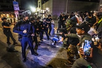 A line of police officers stands on the street at nighttime, holding their batons out, across from a crowd of protesters. In the foreground, a person records on their cellphone.