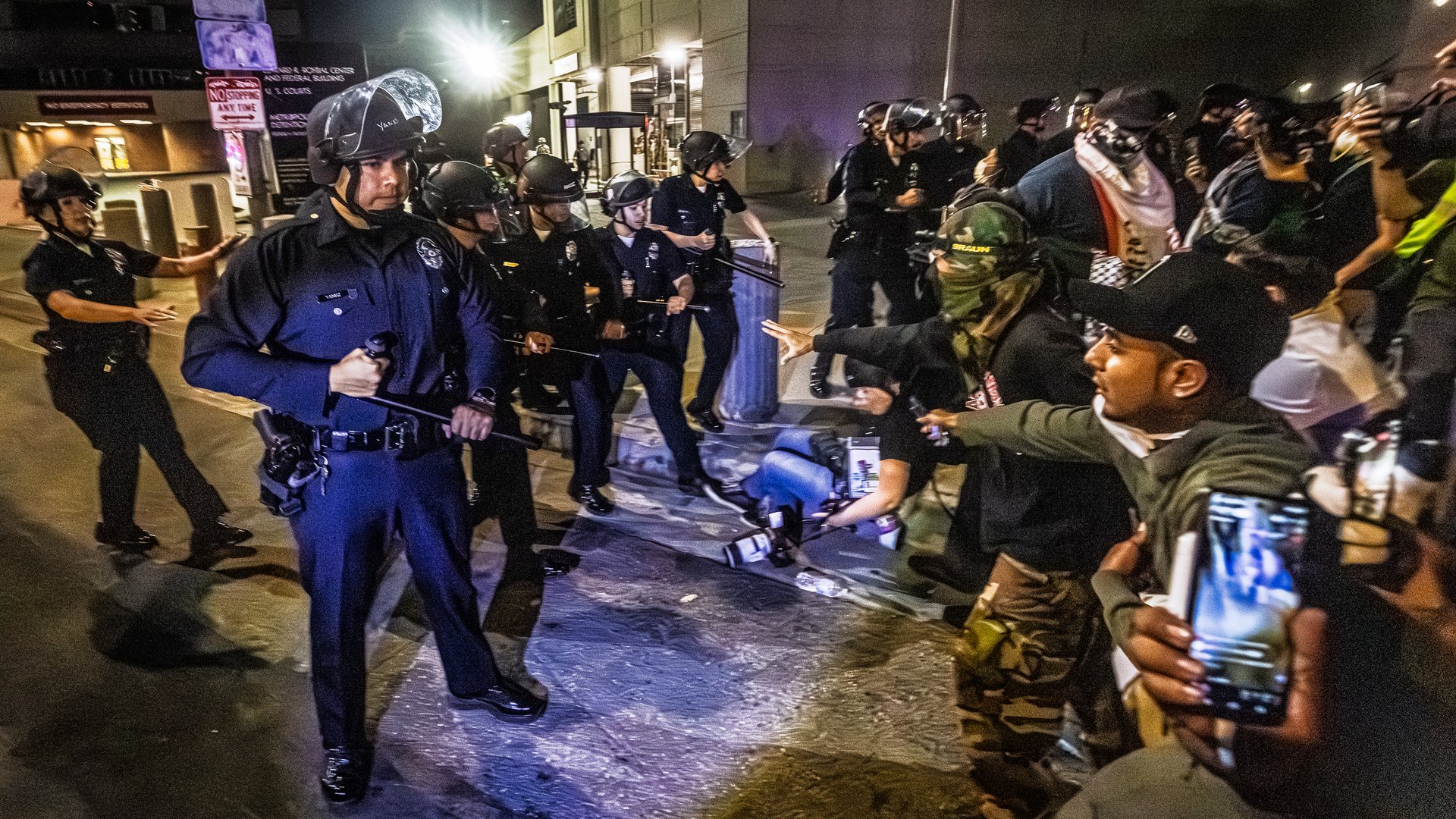 A line of police officers stands on the street at nighttime, holding their batons out, across from a crowd of protesters. In the foreground, a person records on their cellphone.