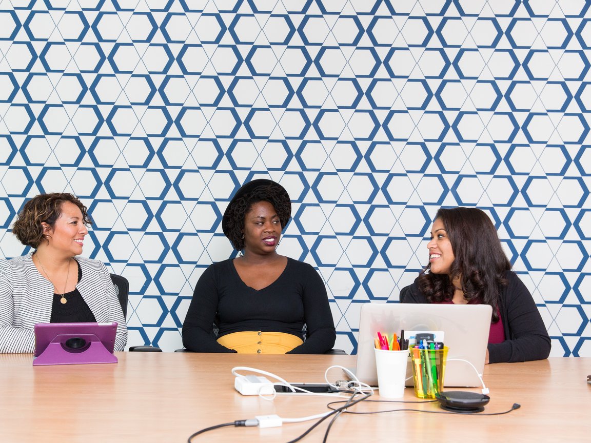 Three women sitting at an office table in front of computers and tablets.