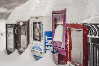 Five newspaper boxes of various colors in a row, covered in deep snow