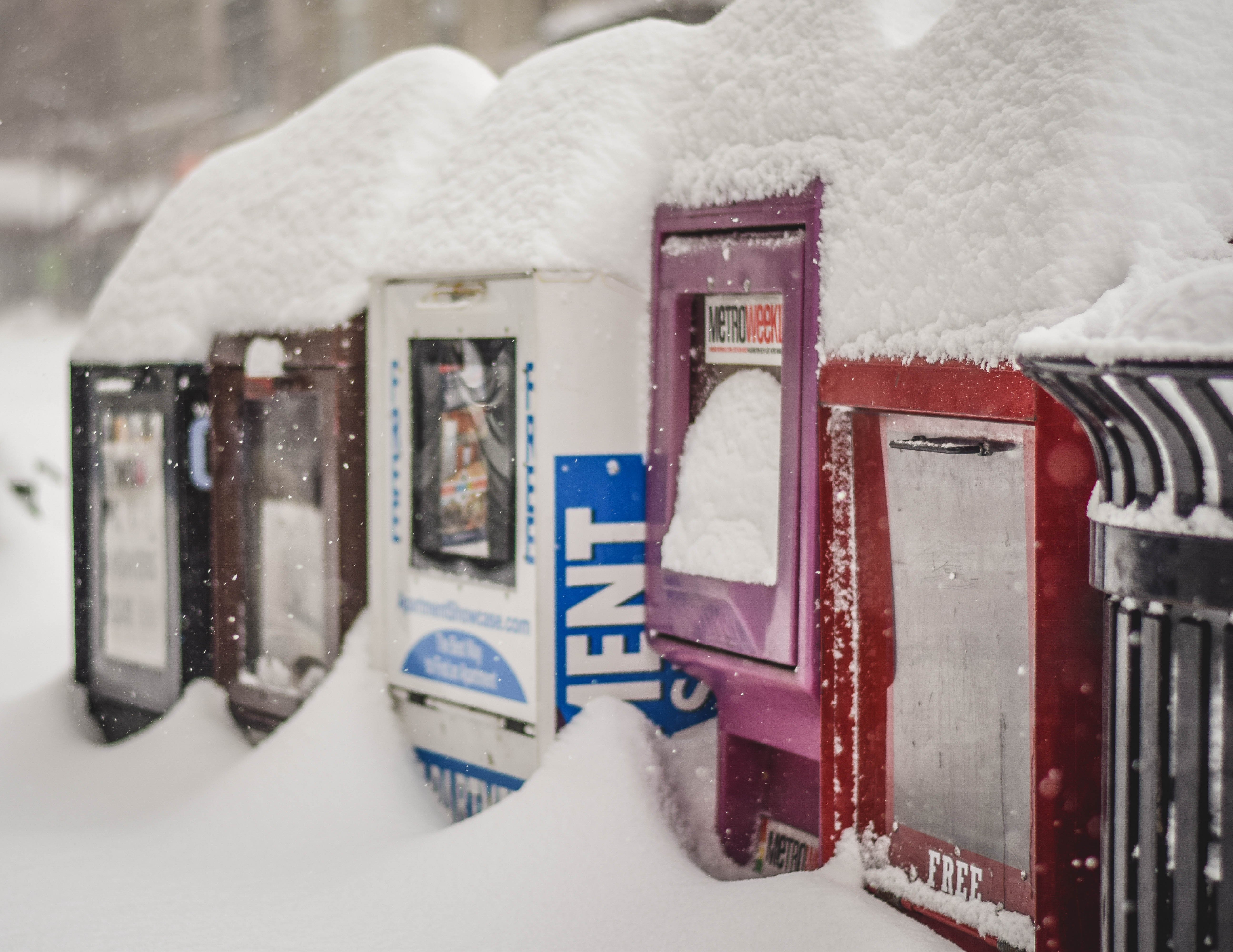 Five newspaper boxes of various colors in a row, covered in deep snow