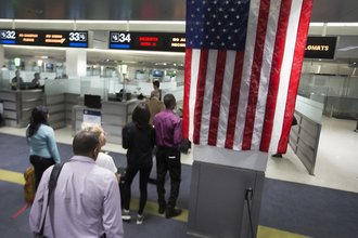 Photo from a United States point of entry with an American flag hanging from the ceiling, as well as a line of travelers leading to an officer screening zone.