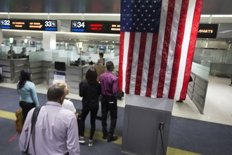 Photo from a United States point of entry with an American flag hanging from the ceiling, as well as a line of travelers leading to an officer screening zone.