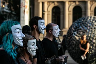 Four protesters wearing Guy Fawkes masks in front of a neoclassical building with a modern sculpture and a blurry onlooker in front of it.