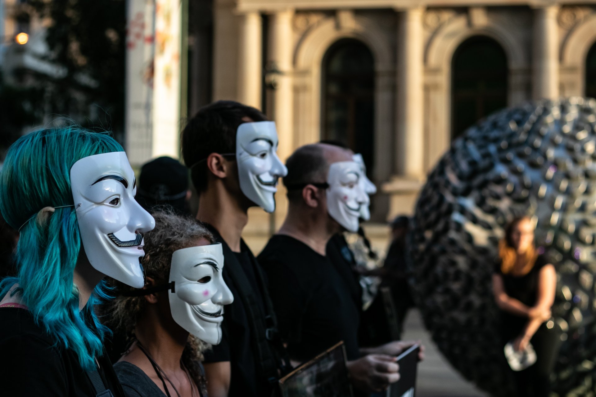Four protesters wearing Guy Fawkes masks in front of a neoclassical building with a modern sculpture and a blurry onlooker in front of it.