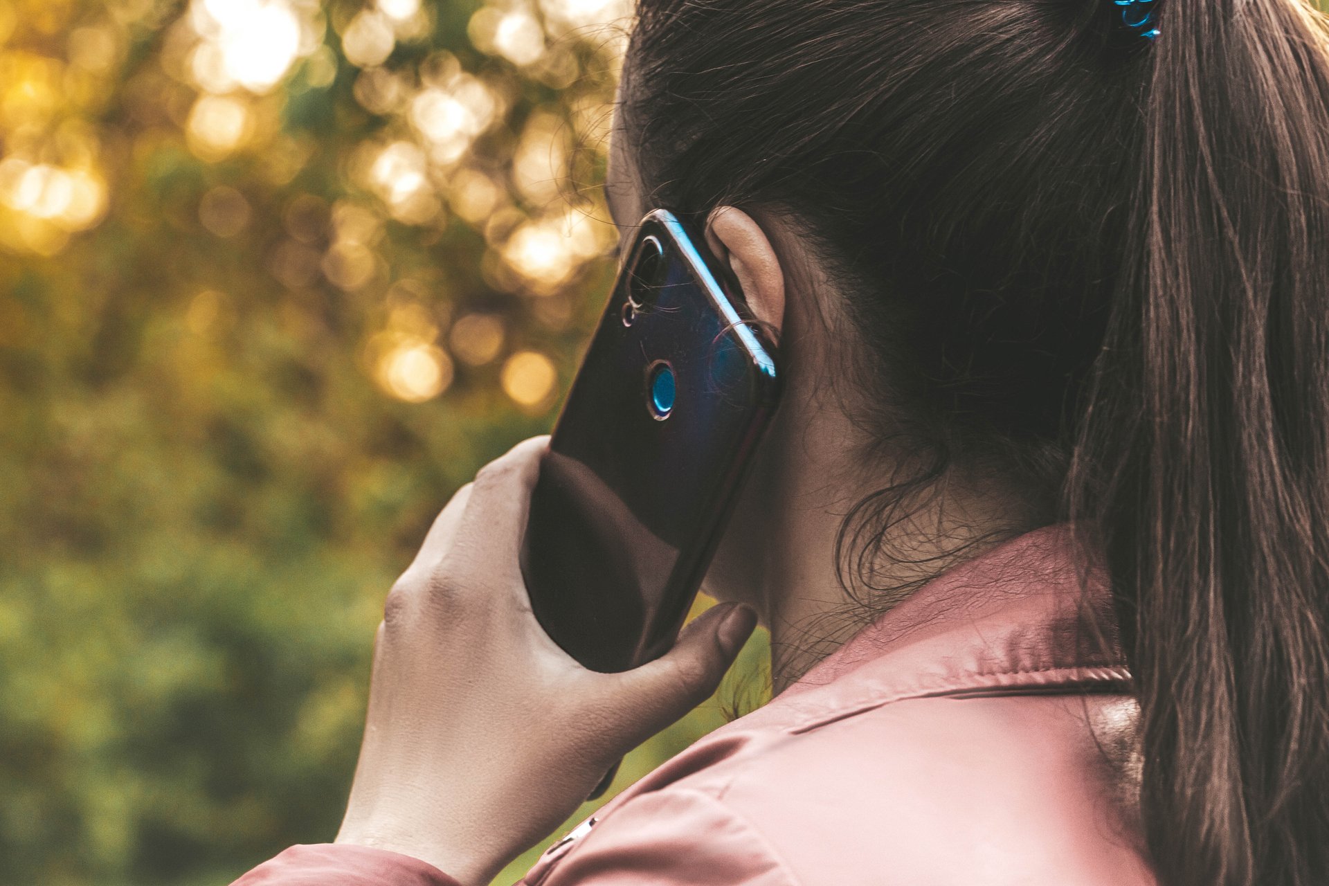 The back of a woman's head, showing her pony tail, pink jacket, and phone held up to her hear