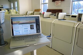 An open laptop sits on a table in a laundromat, in front of several washing mashines