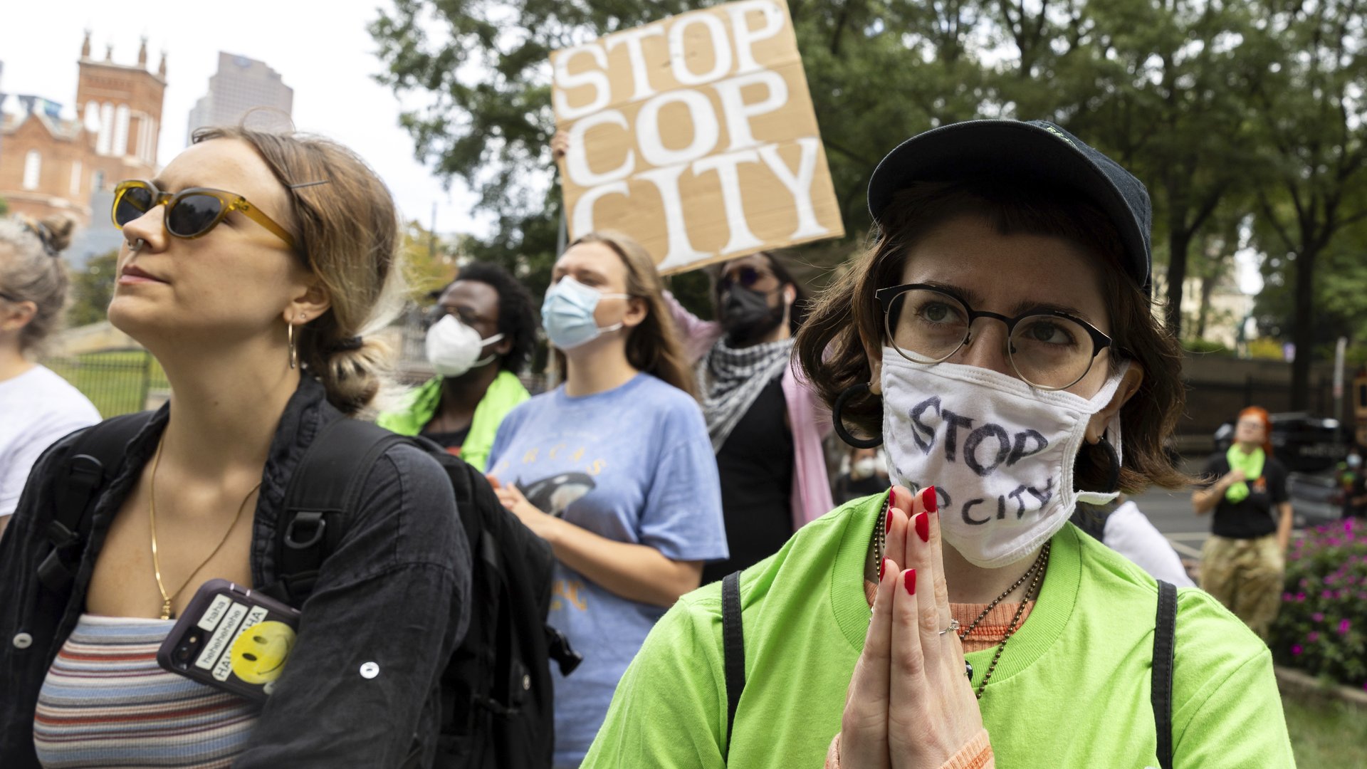 A masked protester in the foreground holds their hands together while protesters in the background hold signs saying things like Stop Cop City.