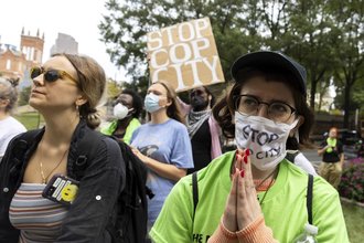 A masked protester in the foreground holds their hands together while protesters in the background hold signs saying things like Stop Cop City.