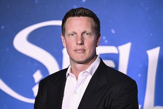 David Ellison, wearing a dark suit jacket and light shirt, stands facing the camera and staring, deadeyed, in front of a blue background with white lettering.