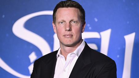 David Ellison, wearing a dark suit jacket and light shirt, stands facing the camera and staring, deadeyed, in front of a blue background with white lettering.