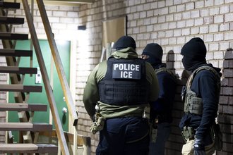 Three officers stand in a stairwell, wearing vests and balaclavas. The words “POLICE ICE” appear on the vest of one officer with his back toward the camera.