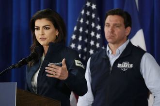 Florida first lady Casey DeSantis stands at a podium speaking into a microphone, while her husband, Florida Gov. Ron DeSantis, looks on, with an American flag in the background.