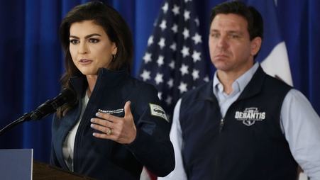 Florida first lady Casey DeSantis stands at a podium speaking into a microphone, while her husband, Florida Gov. Ron DeSantis, looks on, with an American flag in the background.