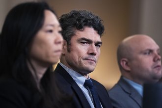 Joe Kent, in focus and wearing a dark suit and tie, sits facing the camera between two other people.