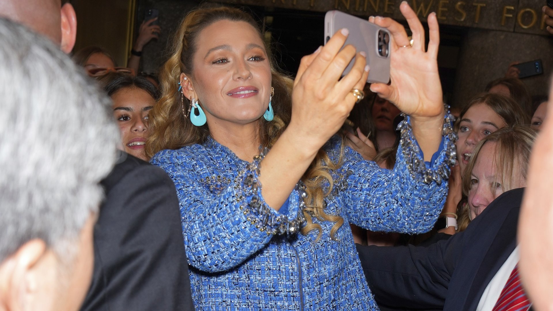 A midst of a blonde woman in a blue jacket taking a photo with an iPhone while surrounded by other people.