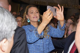A midst of a blonde woman in a blue jacket taking a photo with an iPhone while surrounded by other people.