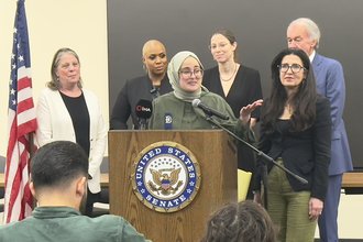 Tufts student Rümeysa Öztürk speaks from a podium with a U.S. Senate logo, surrounded by a group of people.