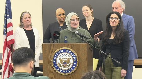 Tufts student Rümeysa Öztürk speaks from a podium with a U.S. Senate logo, surrounded by a group of people.