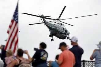 President Donald Trump’s helicopter flies away, with blurred images of people and an American flag in the foreground.