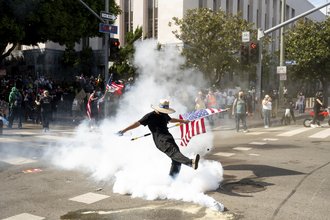 A photograph of a protester wearing a hat and holding the American flag, dressed in all black, in a plume of tear gas fumes seen kicking the tear gas canister away.