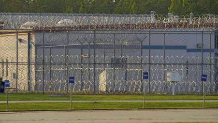 A barbed-wire fence surrounding a detention facility, with signs reading “DHS” outside.
