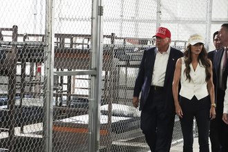 Trump and Kristi Noel walking on a tour of Alligator Alcatraz in front of a chain-link fence with bunk beds behind the fence.