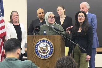 Rümeysa Öztürk, wearing a light green hijab and green top, speaks into a microphone at a podium labeled “United States Senate” and gestures toward the audience. She is flanked by five supporters.
