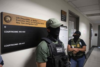 A masked federal agent stands outside a courtroom wearing a bulletproof vest, with another similarly dressed agent in the background.