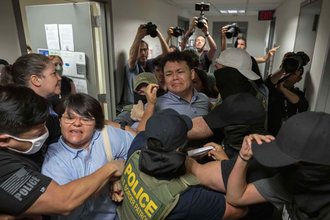 A man in a blue shirt with an anguished expression is held by other men wearing law enforcement uniforms and masks, while a woman in a blue shirt is pulled away by another