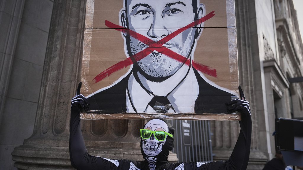 A person wearing a skeleton costume and green sunglasses holds up a sign with a drawing of Jimmy Kimmel’s face and a red X over his mouth.