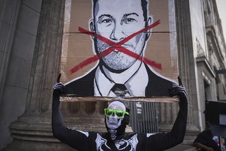 A person wearing a skeleton costume and green sunglasses holds up a sign with a drawing of Jimmy Kimmel’s face and a red X over his mouth.