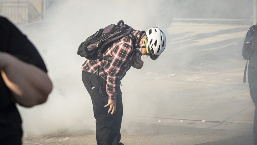 A man wearing a bicycle helmet and backpack stands outside, bent at the waist with one hand on his leg and the other pulling down a mask from his mouth, with a cloud of gas around him.