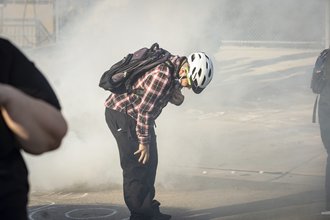 A man wearing a bicycle helmet and backpack stands outside, bent at the waist with one hand on his leg and the other pulling down a mask from his mouth, with a cloud of gas around him.