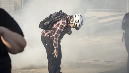 A man wearing a bicycle helmet and backpack stands outside, bent at the waist with one hand on his leg and the other pulling down a mask from his mouth, with a cloud of gas around him.