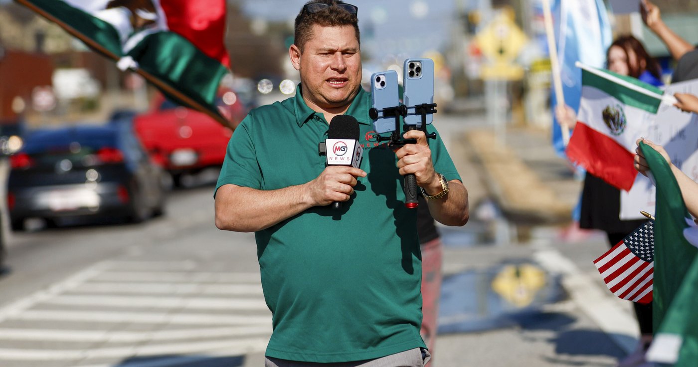 A man is standing in the street livestreaming with two phones and a microphone
