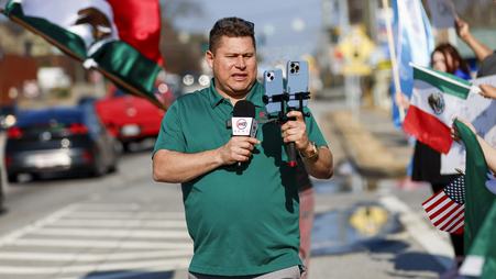 Journalist Mario Guevara, in a green shirt, stands in the street livestreaming with two phones and a microphone