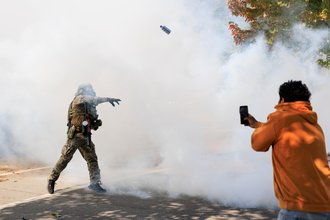 A photograph of a law enforcement officer in military-style gear throwing a tear gas canister at an onlooker who is recording on their phone. Tear gas or smoke is covering nearly the entire space within the frame.