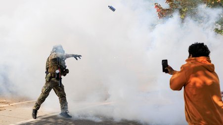 A photograph of a law enforcement officer in military-style gear throwing a tear gas canister at an onlooker who is recording on their phone. Tear gas or smoke is covering nearly the entire space within the frame.