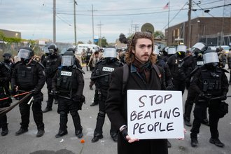 A man holds a white sign that says "Stop beating people" in the foreground with a line of law enforcement in full riot gear in the background