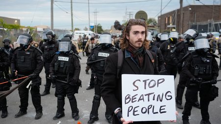 A man holds a white sign that says "Stop beating people" in the foreground with a line of law enforcement in full riot gear in the background