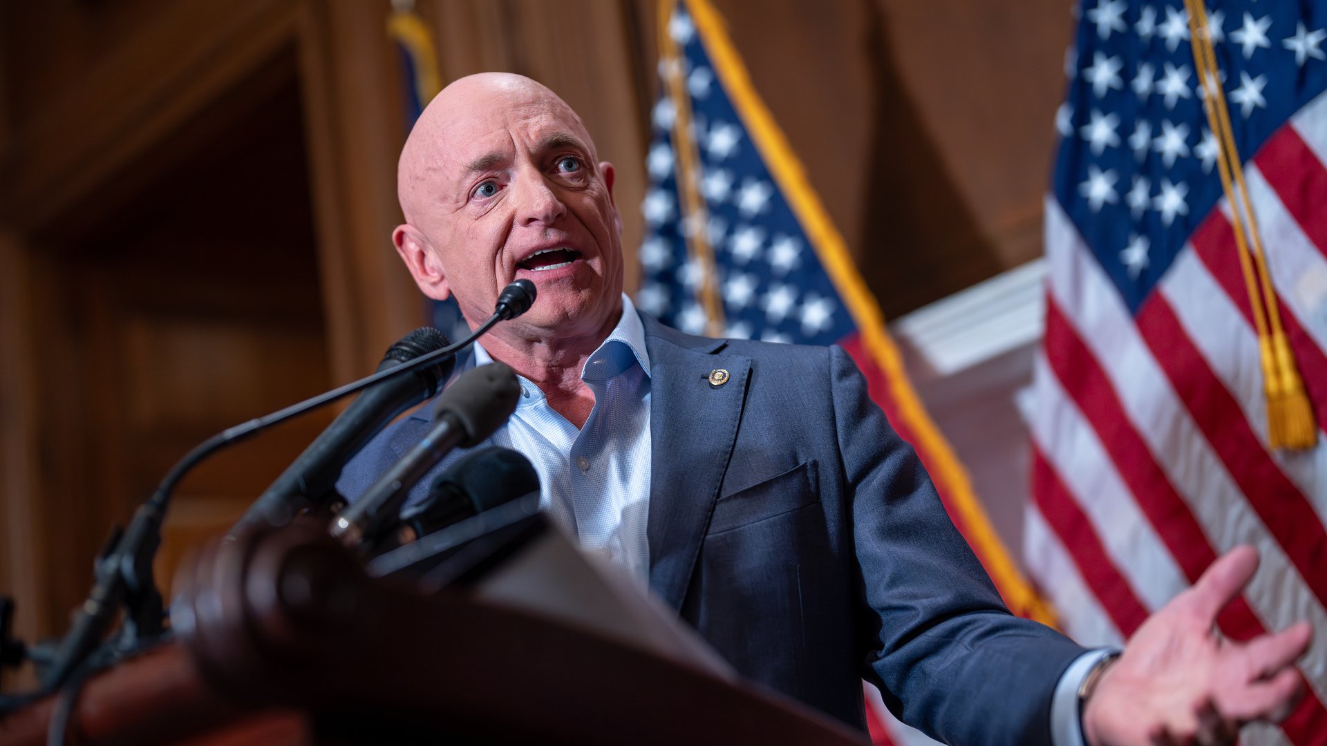 A mid-shot of a bald white man in a suit, Senator Mark Kelly, emoting at a podium with two American flags in the background.