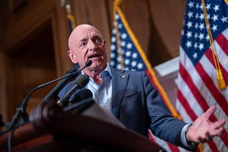 A mid-shot of a bald white man in a suit, Senator Mark Kelly, emoting at a podium with two American flags in the background.